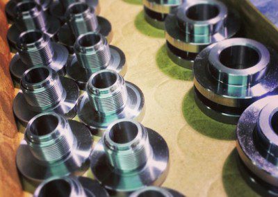 a collection of machined metallic parts with a silvery finish, arranged in rows inside a cardboard box. Each part has a polished, cylindrical shape with a flanged base and a threaded top section, indicative of their use in industrial or mechanical settings.