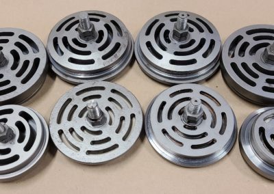a collection of disc-shaped metal components with intricate, curving cut-out patterns on their faces, arranged on a beige surface. The center of each disc features a protruding hub with a knurled texture, suggesting these could be rotary elements, such as impellers or part of a mechanical assembly that requires balanced, symmetrical designs for functionality.